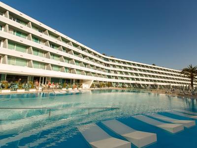 Large hotel building with a long, curved pool and sun loungers in the water under clear sky.