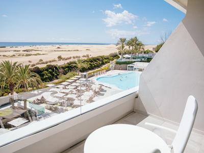 Balcon avec table et chaises offrant une vue sur la piscine, les palmiers et la plage sous un ciel bleu.