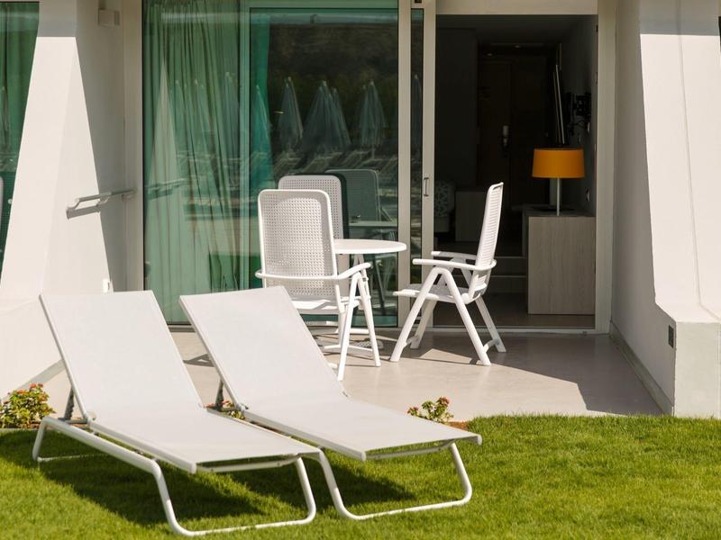 Terrasse d'hôtel avec chaises longues blanches sur pelouse verte devant une table et chaises sous un porche.