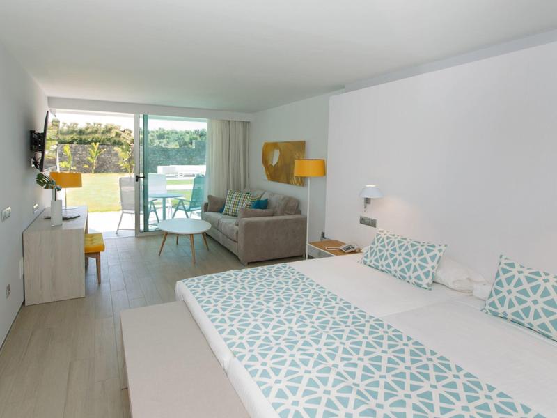 Chambre d'hôtel lumineuse avec lit double, canapé, bureau et vue sur balcon verdoyant.