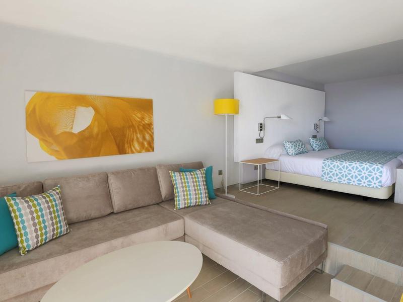 Chambre d'hôtel moderne avec canapé, lit double, lampe jaune et décoration murale jaune.