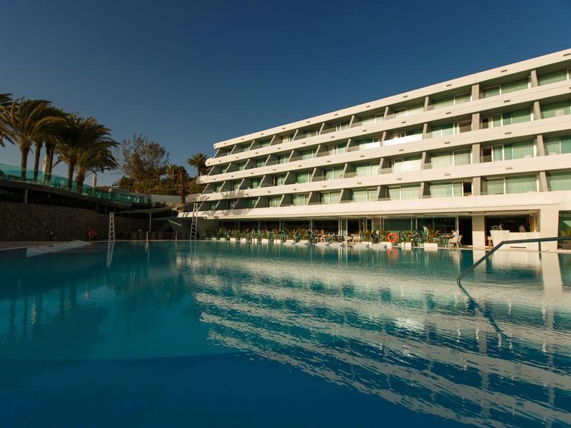 Hôtel moderne avec plusieurs étages et grande piscine extérieure sous un ciel clair.
