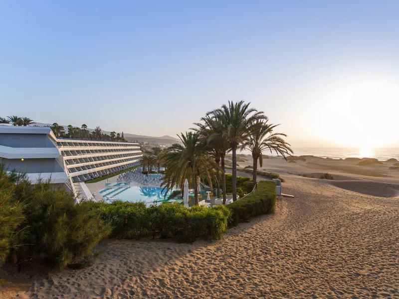 Hôtel moderne avec piscine, palmiers et plage de sable au coucher du soleil.