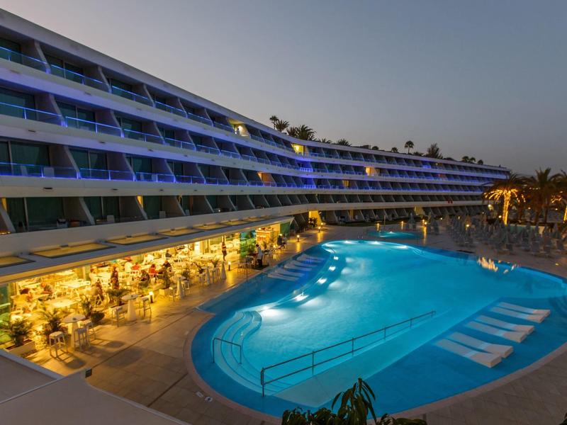 Piscine éclairée en soirée devant un hôtel moderne aux balcons lumineux.