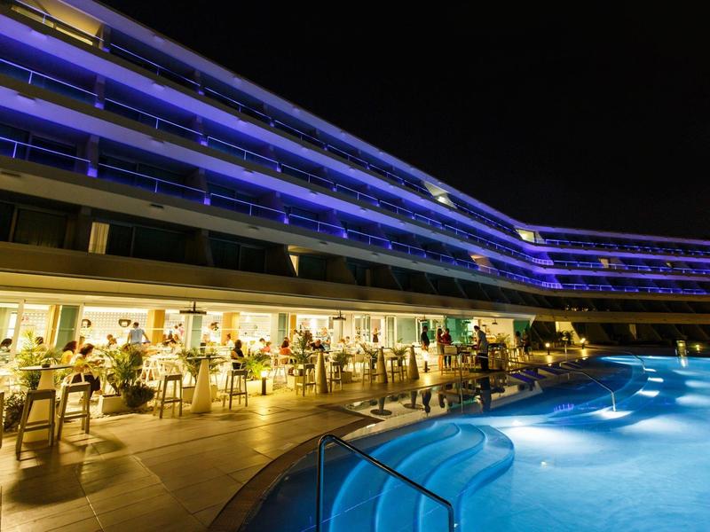 Piscine bleue éclairée la nuit devant un hôtel avec terrasse animée.