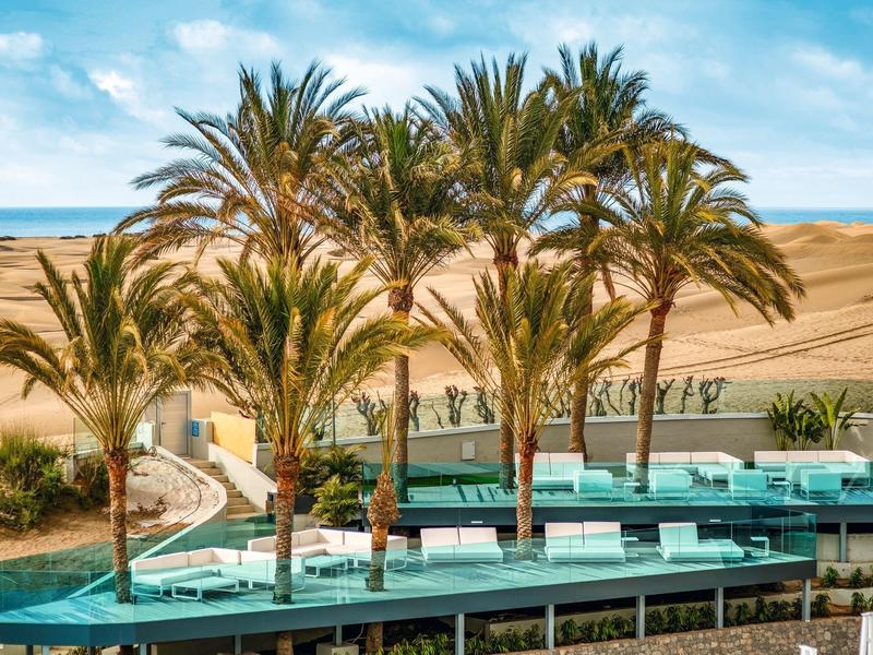 Piscine entourée de palmiers avec vue sur un désert et ciel bleu nuageux.