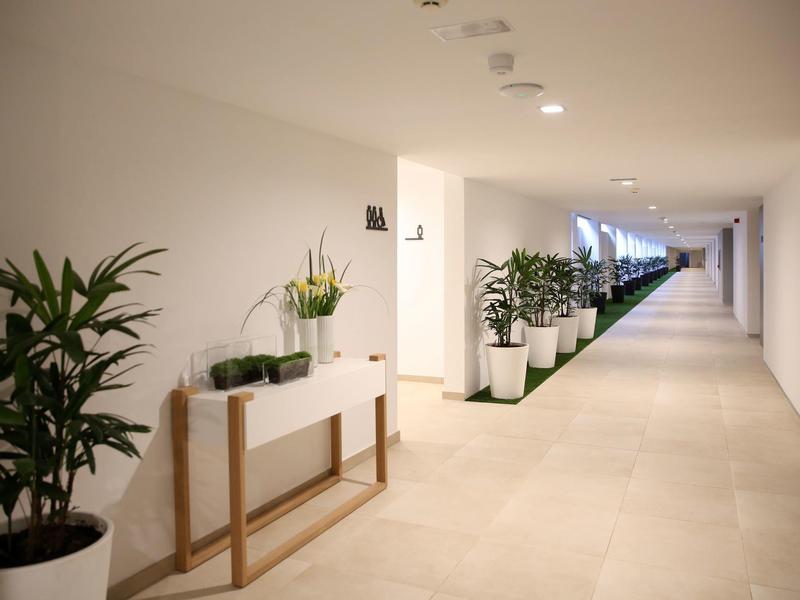 Couloir d'hôtel lumineux avec plantes en pot alignées sur un mur blanc et sol carrelé clair.