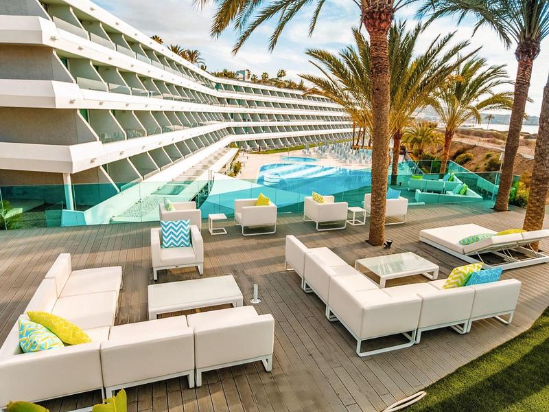 Terrasse d'hôtel avec canapés blancs, piscine et palmiers sous un ciel clair.