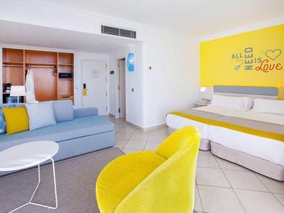 Jasny pokój hotelowy z żółtymi akcentami, łóżkiem podwójnym i częścią wypoczynkową z sofą i krzesłem.