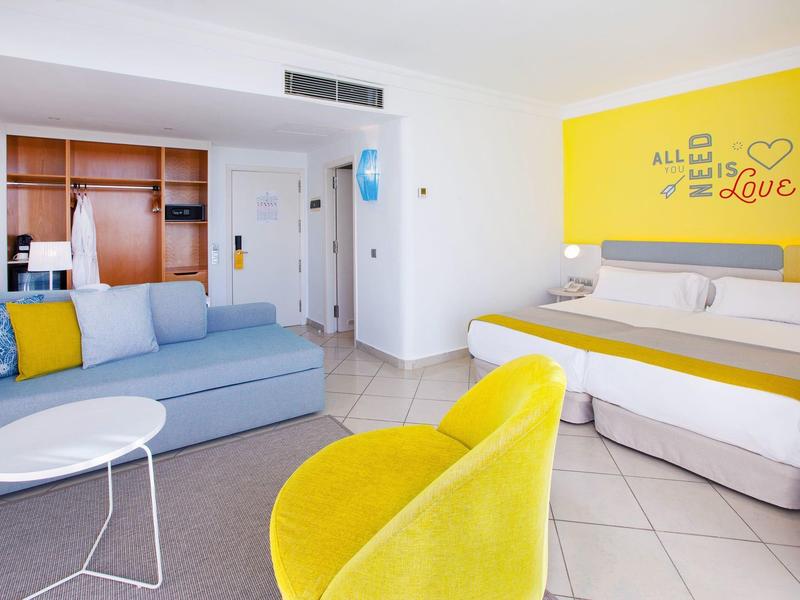 Jasny pokój hotelowy z żółtymi akcentami, łóżkiem podwójnym i częścią wypoczynkową z sofą i krzesłem.