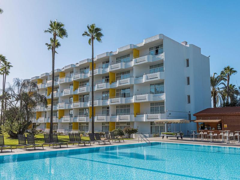 Nowoczesny budynek hotelowy z pokojami z balkonem, palmami i dużym basenem w słoneczną pogodę.