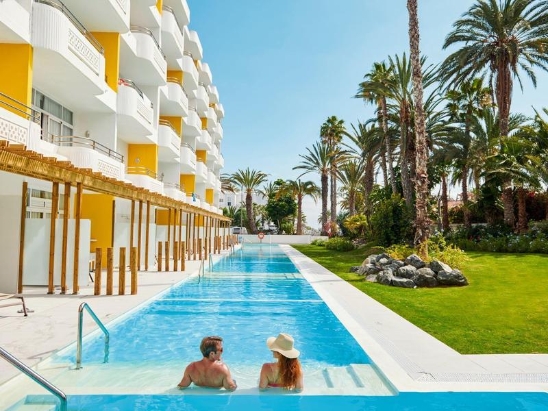 Dwie osoby w basenie hotelowym z białymi budynkami i palmami w słoneczny dzień