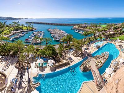 Blick auf eine Hotelanlage mit Pool, Liegestühlen und einem Yachthafen am Meer bei klarem Himmel.