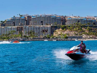 Mehrere Personen auf Jetskis und einem Schlauchboot nahe einer Küstenstadt bei Sonnenschein.