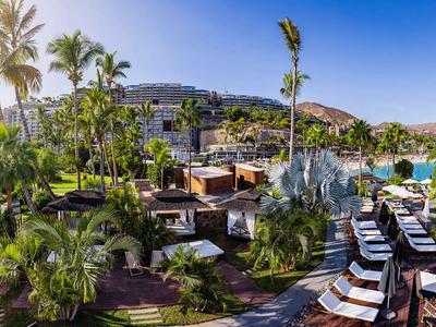 Hotelanlage mit Pool, Liegestühlen und Palmen unter blauem Himmel in sonniger Landschaft.