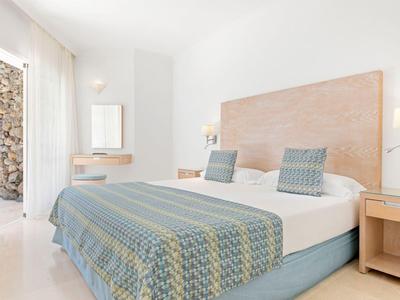 Helles Schlafzimmer mit Doppelbett, blauer Tagesdecke und großen Fenstern mit Blick nach draußen.