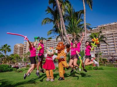 Kinder und Maskottchen springen auf einer grünen Wiese vor einem Hotel mit Palmen.