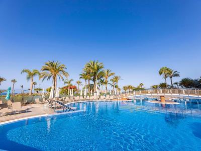 Runder Außenpool mit Palmen und Sonnenliegen unter klarem blauem Himmel.