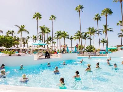Großer Pool mit vielen Menschen und Palmen im Hintergrund in einem Resort.