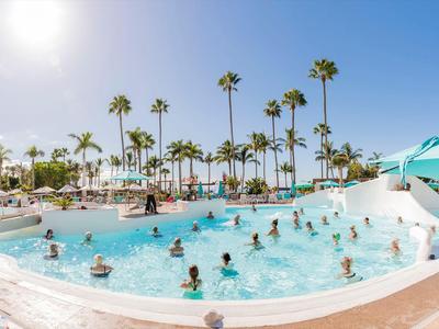 Belebter Pool mit Menschen und Palmen bei sonnigem Himmel in einem Resort.