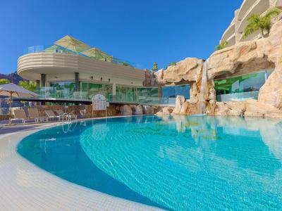 Moderner Hotelpool mit Felsenmotiv und Sonnenterrasse unter klarem blauem Himmel.