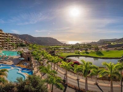 Panoramablick auf ein Hotelresort mit Pool, Palmen und Bergen im Hintergrund bei Sonnenlicht.