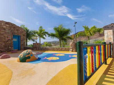 Bunter Outdoor-Spielplatz mit Sand, Kletterelementen und Palmen im Hintergrund.