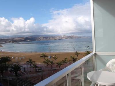 Balkon mit weißem Stuhl und Glasgeländer blickt auf Strand, Meer und bewölkten Himmel.