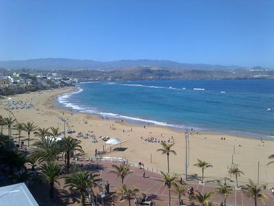 Langer Sandstrand mit blauem Meer, Palmen und Bergen im Hintergrund bei klarem Himmel.