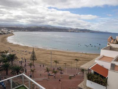 Strand mit goldfarbenem Sand, Palmen und ruhigem Meer unter bewölktem Himmel an einer Küstenstadt.