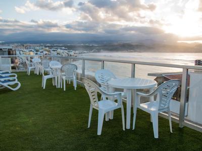 Terrasse mit weißen Plastikstühlen und Tischen, grüner Boden und Sonnenuntergang über dem Meer.
