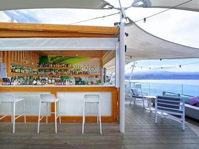 Moderner Außenbarbereich mit weißen Hockern, Holzverkleidung und Blick auf das Meer bei bewölktem Himmel.