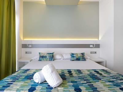 Modernes Schlafzimmer mit weißem Bett, blau-grünem Bettüberwurf, Kissen und grünen Vorhängen.