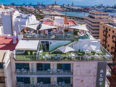 Mehrstöckiges Gebäude mit Dachterrasse, Sonnenschirmen, Pflanzen und Hafen im Hintergrund.