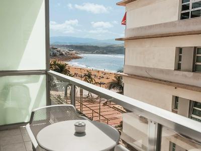Balkon mit runden weißem Tisch, zwei weißen Stühlen und Blick auf Gebäude und Wasser im Hintergrund.
