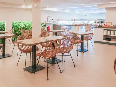 Moderner heller Cafébereich mit Korbstühlen und Tischen auf hellem Boden und Kaminwand.