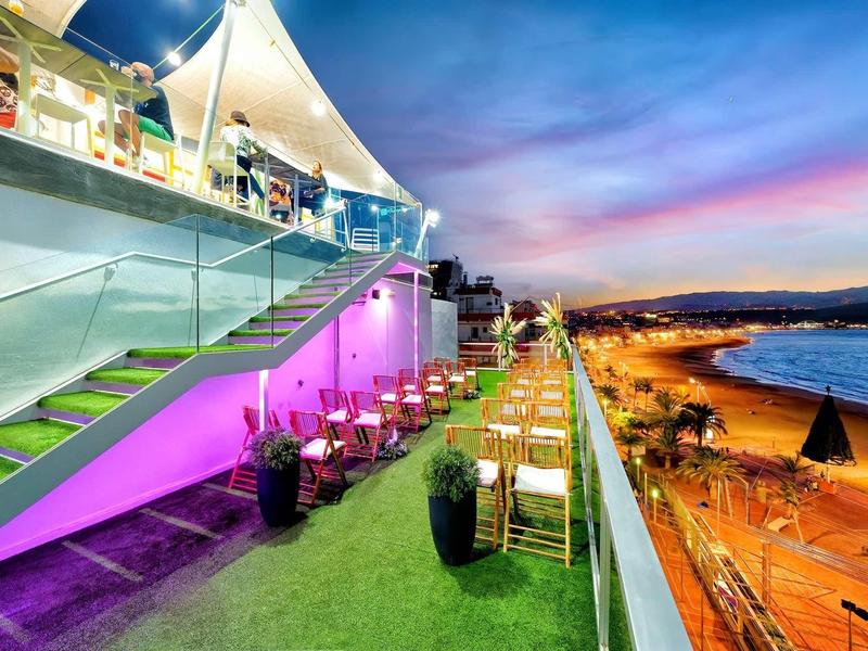 Terrasse mit bunten Lichtern, Tischen und Stühlen neben einem Strand bei Sonnenuntergang.