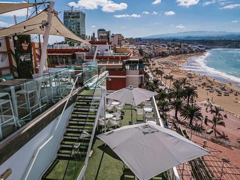 Blick von einer Dachterrasse mit weißen Sonnenschirmen und Glasschutze auf einen belebten Sandstrand.