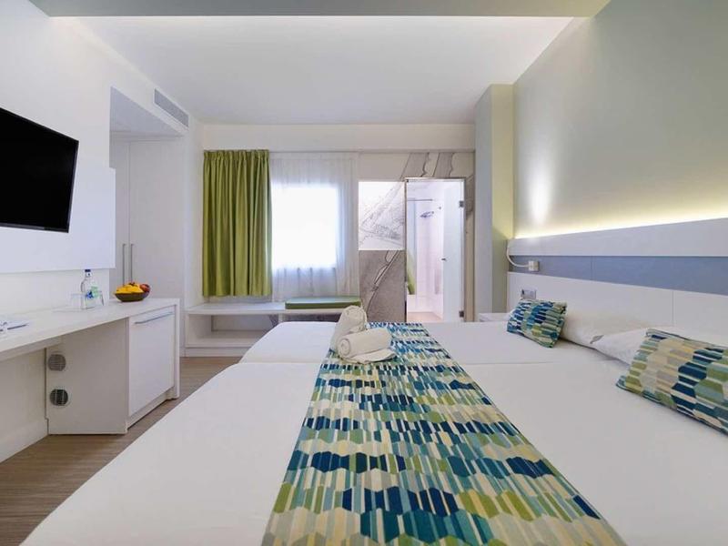 Modernes Hotelzimmer mit großem Bett, bunten Kissen, grünem Vorhang und Flachbildschirm an der Wand.