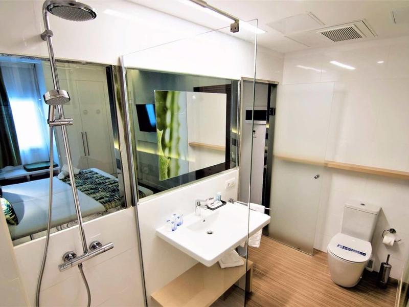 Helles Badezimmer mit großer Spiegelwand, weißem Waschbecken, Dusche und Toilette, helle Fliesen.