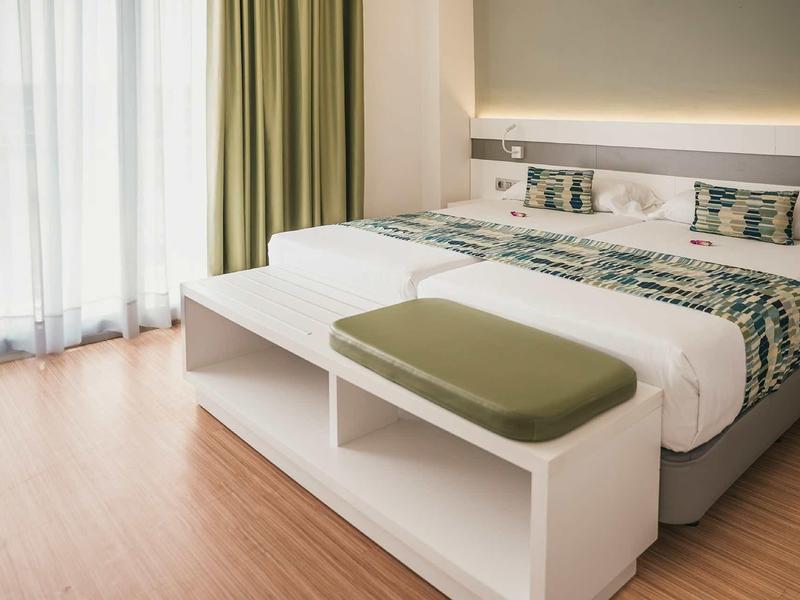 Modernes Schlafzimmer mit großem Doppelbett, weißem Hocker, grünen Vorhängen und Parkettboden.