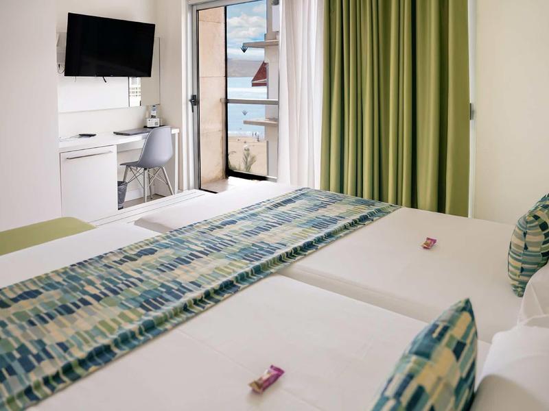 Helles Hotelzimmer mit zwei Betten, grünem Vorhang und Balkon mit Meerblick.