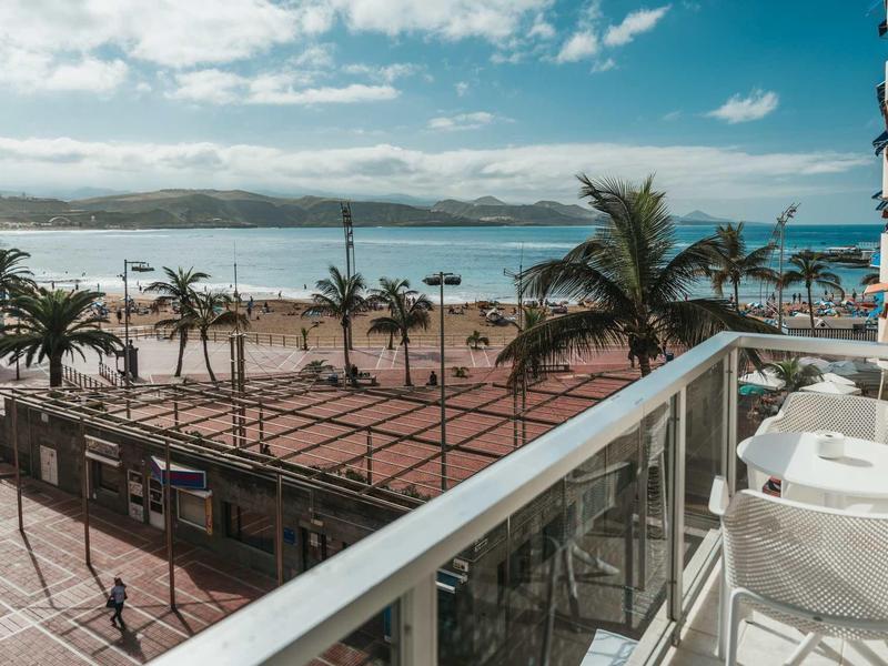 Blick von Balkon mit weißen Stühlen auf Palmen, Strand, Meer und bewölkten Himmel.