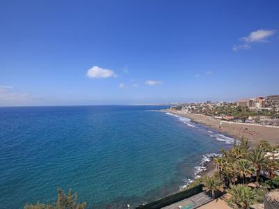 Klares blaues Meer mit Kieselstrand, Palmen und Häusern unter blauem Himmel mit wenigen Wolken.