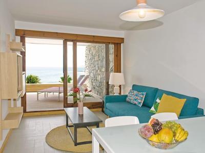 Helles Wohnzimmer mit blauem Sofa, Glas-Couchtisch, runder Teppich und Blick auf Terrasse mit Meer.