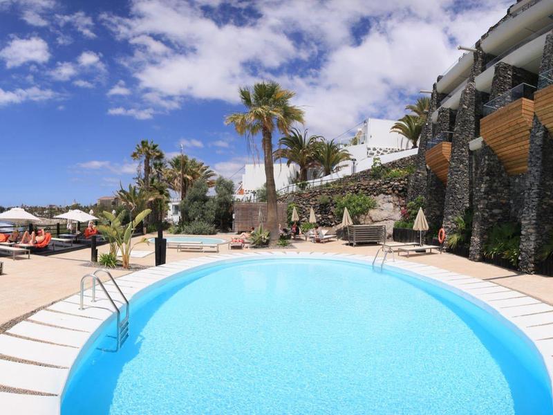 Runder Pool mit klar blauem Wasser neben Sonnenliegen und Palmen unter einem blauen Himmel.