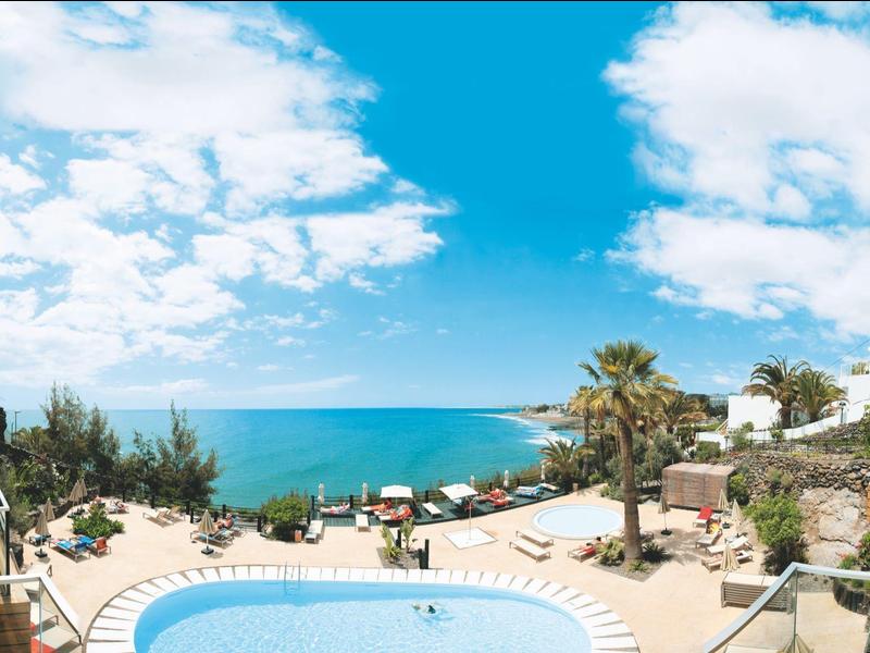 Blick auf einen Außenpool mit Meer im Hintergrund unter blauem Himmel mit Wolken.