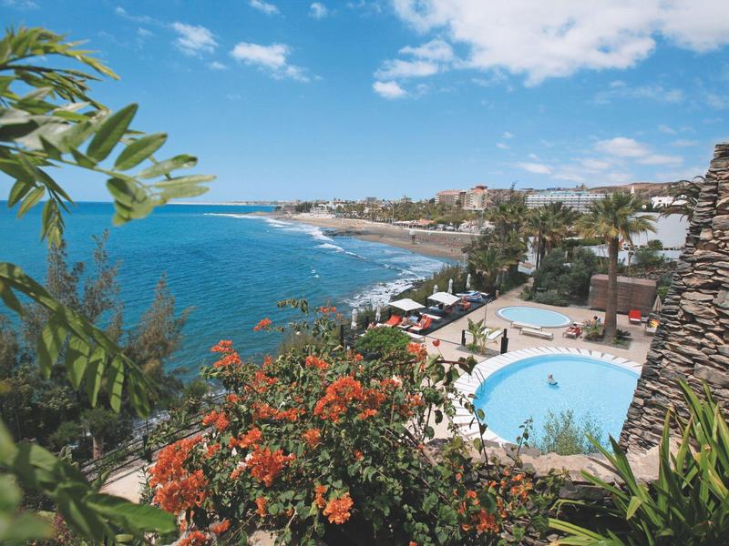 Blick auf ein Küstenhotel mit Pool, Palmen und blühenden Pflanzen unter blauem Himmel.