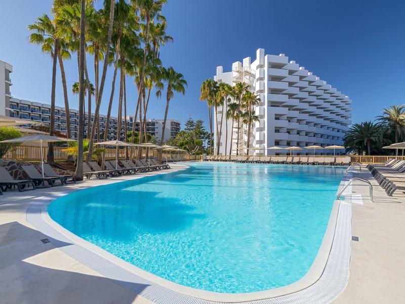 Large outdoor pool with sun loungers and palm trees in front of a modern hotel building under clear sky.
