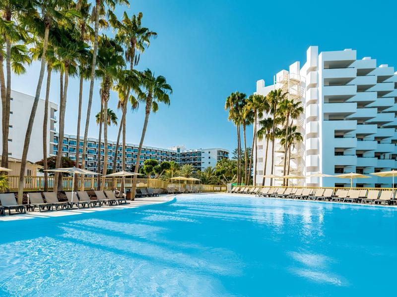 Large hotel pool with lounge chairs, palm trees, and modern building under clear sky.